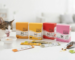 Pet supplements singapore – flat lay of VetSynova cat supplement boxes (KC, LS, RB, SK) and lickable sachets. Happy cat eating from a bowl and kibble scoop nearby on a white surface.